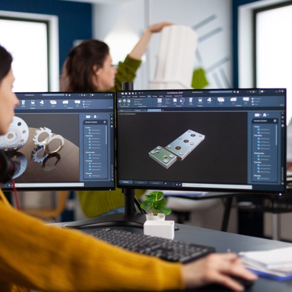 Industrial engineer woman working at pc with two monitors, screens showing CAD software with 3D architecture prototype of gears mechanical piece. Entrepreneur working in creative construction office